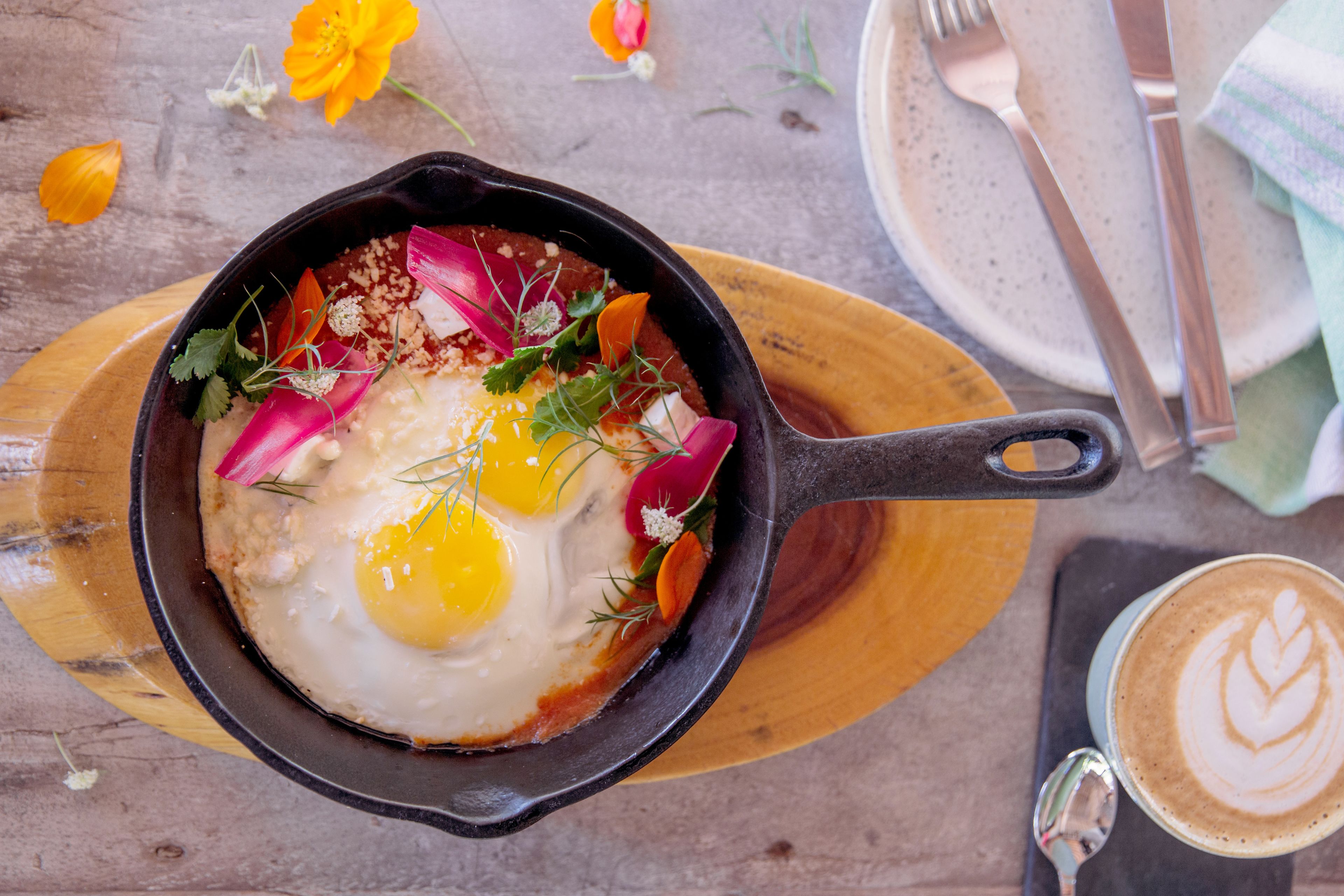 A skillet with eggs and vegetables on a wooden board, a cup of coffee, and a napkin with utensils on a table.