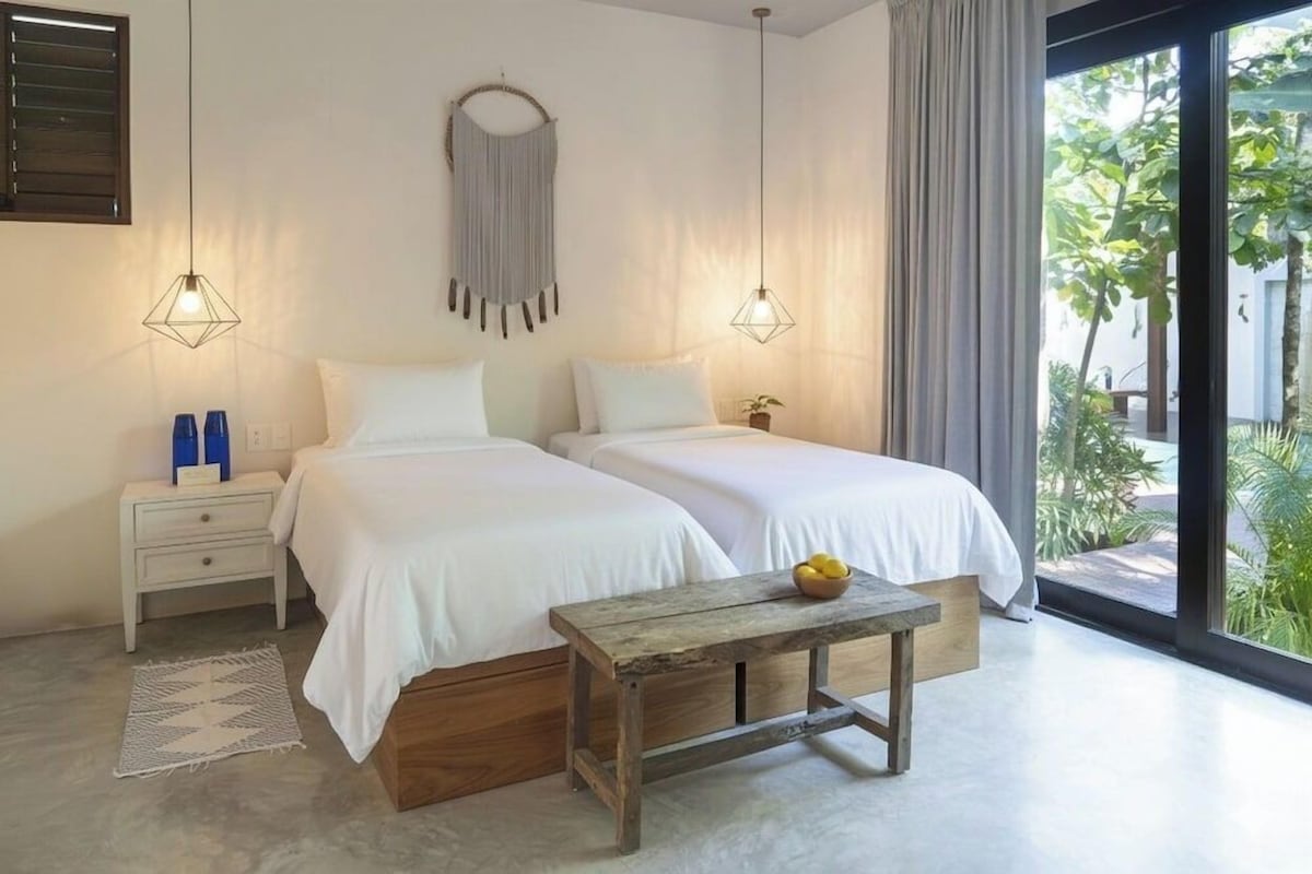 A bedroom with two beds, a nightstand, a wooden bench, and a view of greenery outside.