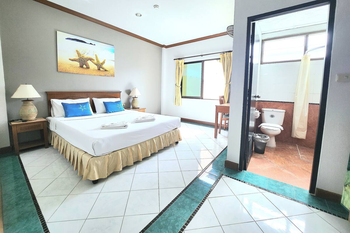 A hotel room with a large bed, a desk, a chair, a window with curtains, a bathroom with a toilet and sink, and a door leading outside.