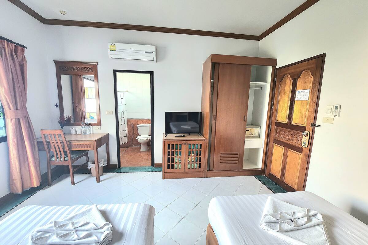 A hotel room with two beds, a TV, a wardrobe, a bathroom visible through a door, and a dining table with chairs.