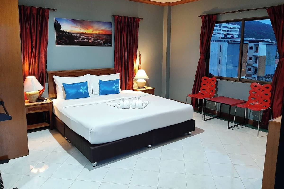 A hotel room with a bed, two bedside lamps, a window with a view of buildings, and a red chair by the window.