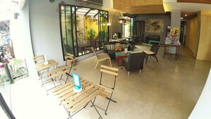 Una cafetería moderna con mesas y sillas de madera, un amplio ventanal con vista a la calle y un rincón acogedor con sofá y un osito de peluche.