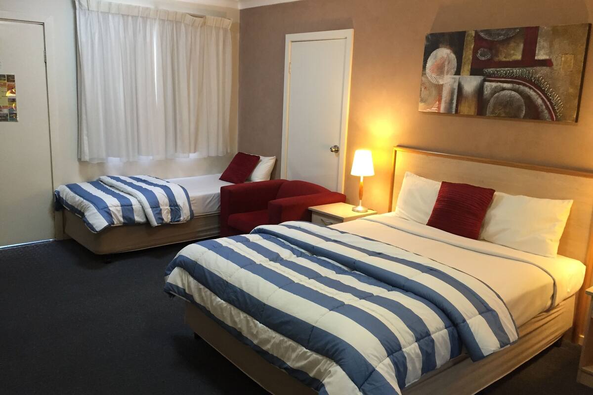 A hotel room with two beds, a red chair, a nightstand with a lamp, and a painting on the wall.