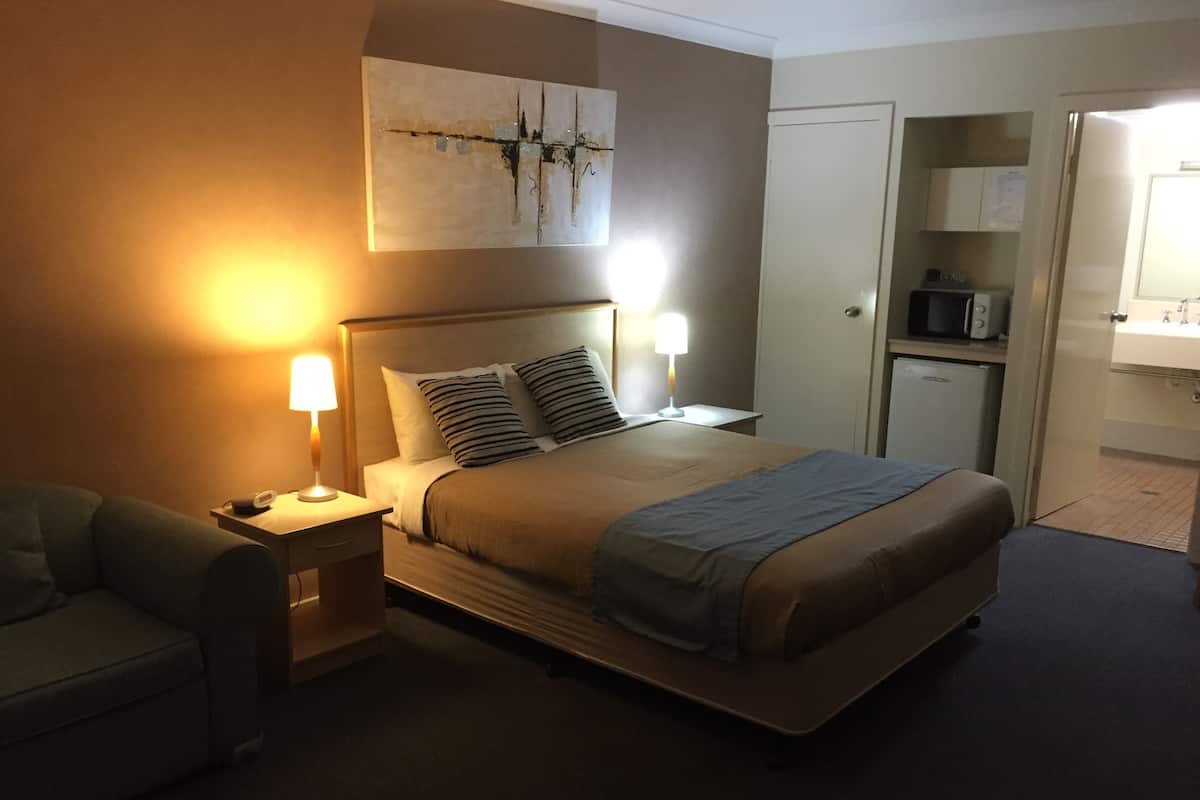 A hotel room with a bed, bedside tables, a lamp, a painting on the wall, and a kitchenette area.