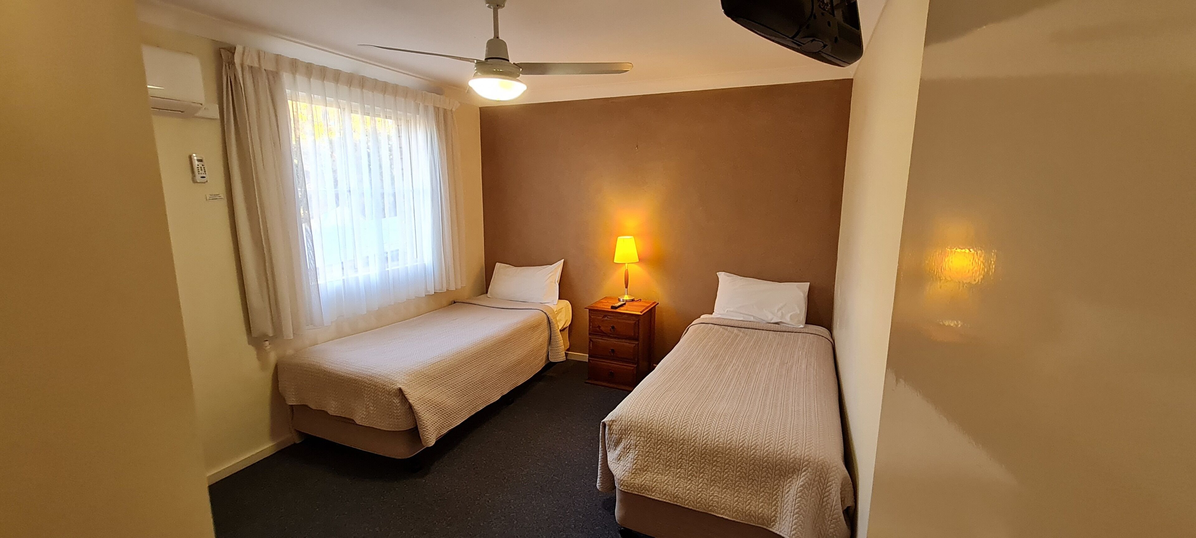 A small hotel room with two single beds, a bedside table with a lamp, a ceiling fan, and an air conditioning unit.
