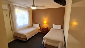 A small hotel room with two single beds, a bedside table with a lamp, a ceiling fan, and an air conditioning unit.