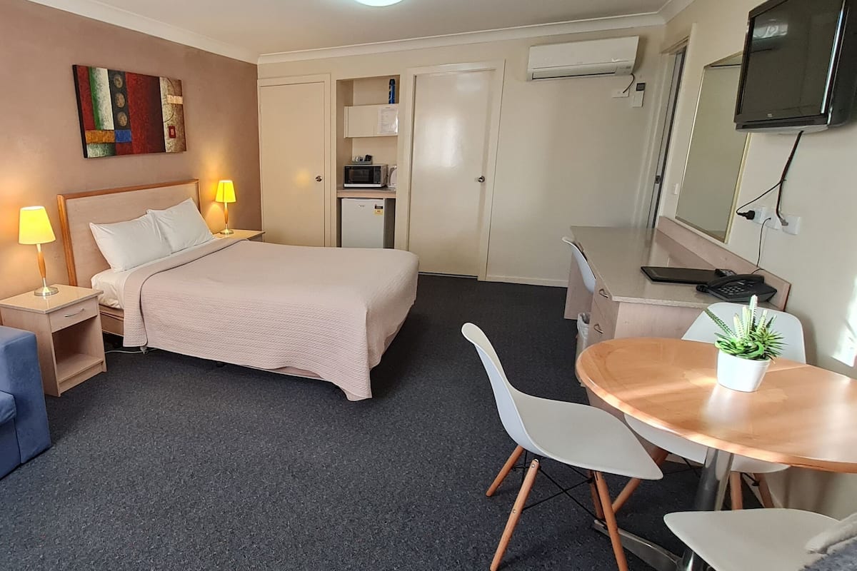 A hotel room with a bed, a blue sofa, a desk with a chair, a small kitchenette, and a TV.
