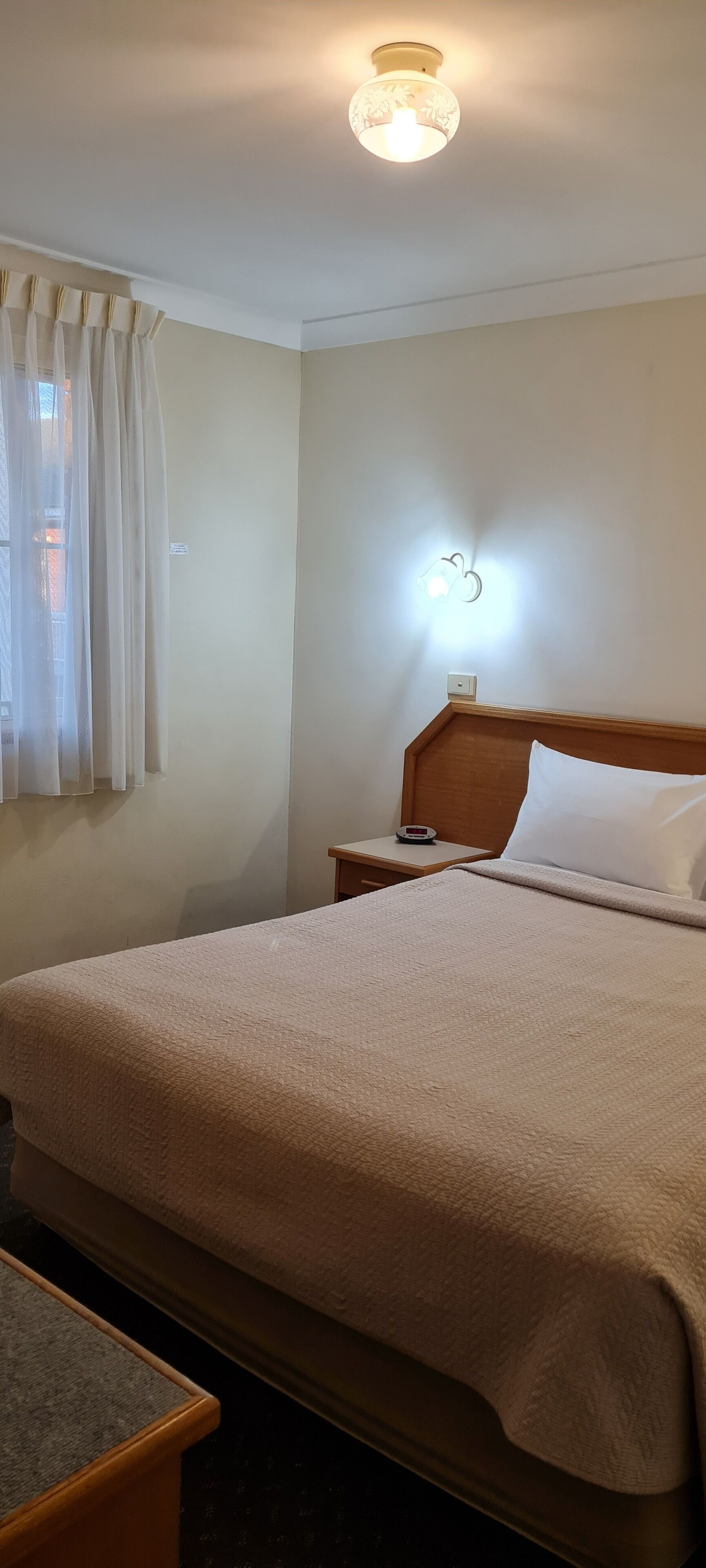 Una habitación de hotel con cama, mesita de noche, lámpara y ventana con cortinas.