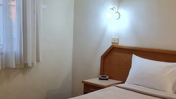 Una habitación de hotel con cama, mesita de noche, lámpara y ventana con cortinas.