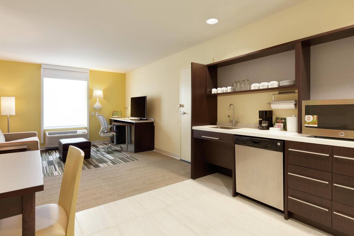 Une chambre d’hôtel moderne dotée d’un coin cuisine, d’une table à manger avec des chaises, d’un canapé, d’une télévision et d’une fenêtre avec des stores.