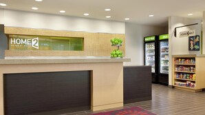 A hotel reception desk with a 'HOME 2' sign, a fruit display, and a vending machine. - Home2 Suites by Hilton Seattle Airport (Tukwila)
