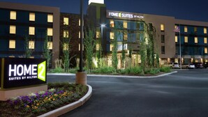 A hotel entrance with a sign reading 'HOME 2 SUITES BY HILTON,' a landscaped area with flowers, and a building with illuminated windows and a flag. - Home2 Suites by Hilton Seattle Airport (Tukwila)