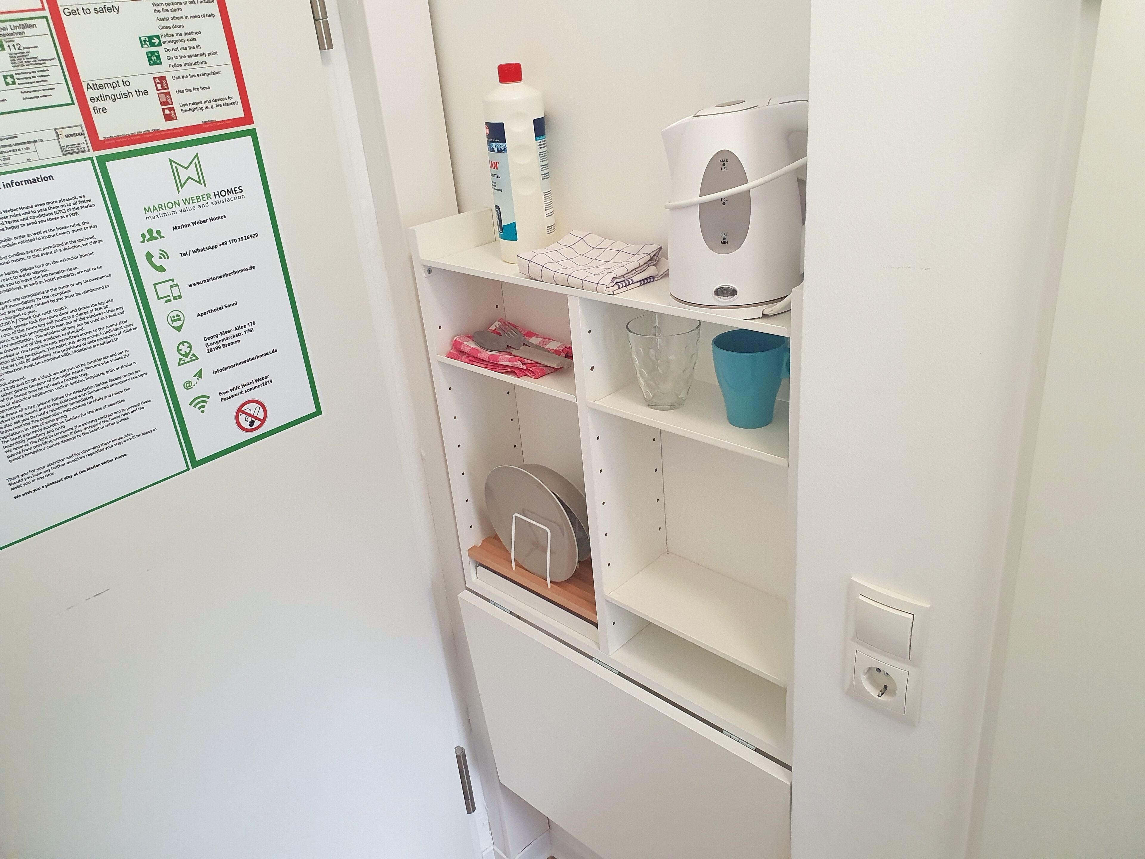 A hotel room with a white wall displaying safety instructions and a small shelf with a bottle, a kettle, and a blue cup.