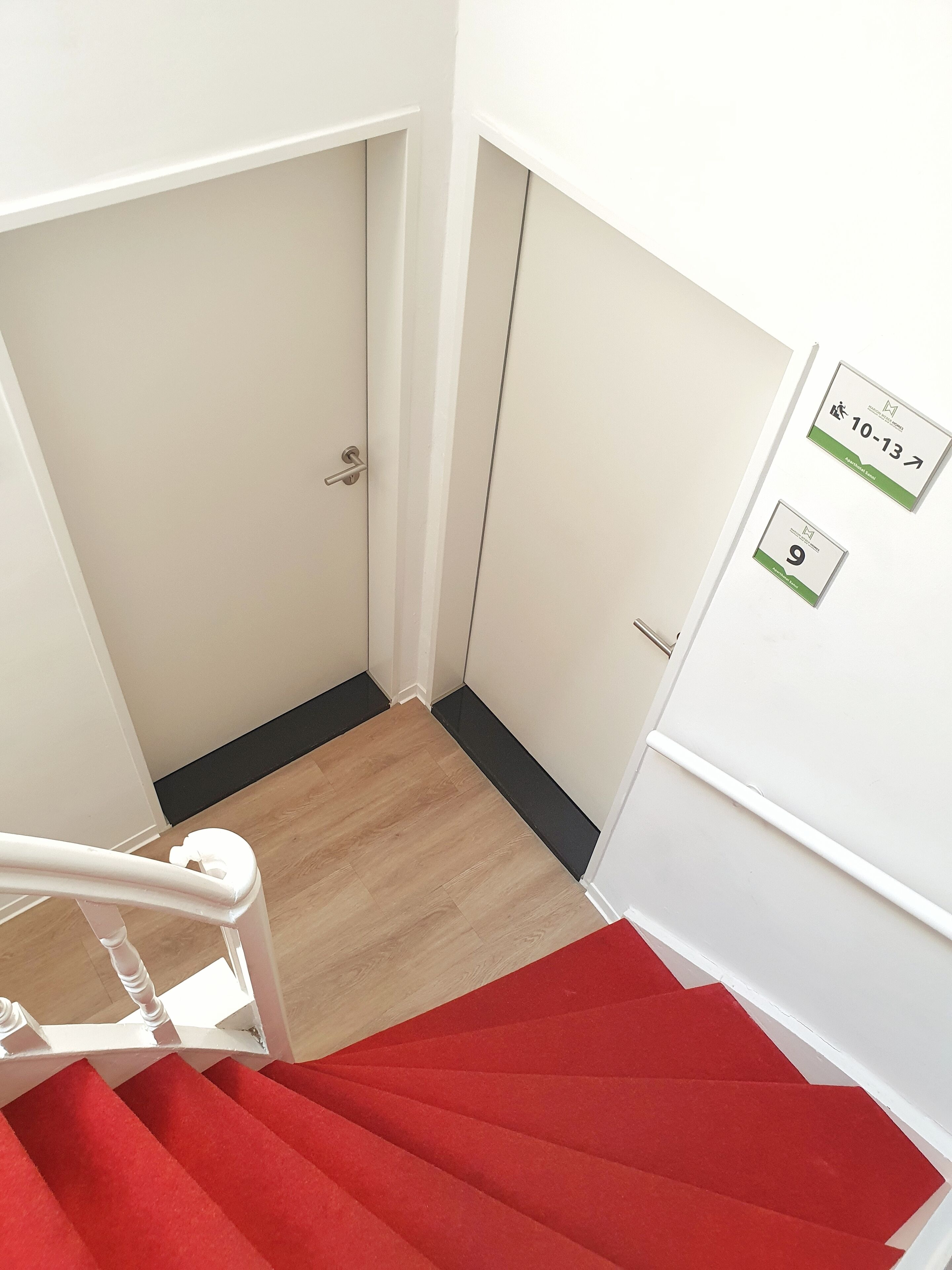 Eine Treppe mit roten Teppichstufen führt zu einer weißen Tür mit Klingel. Zwei Schilder an der Wand zeigen die Zimmernummern 9 und 10–13.