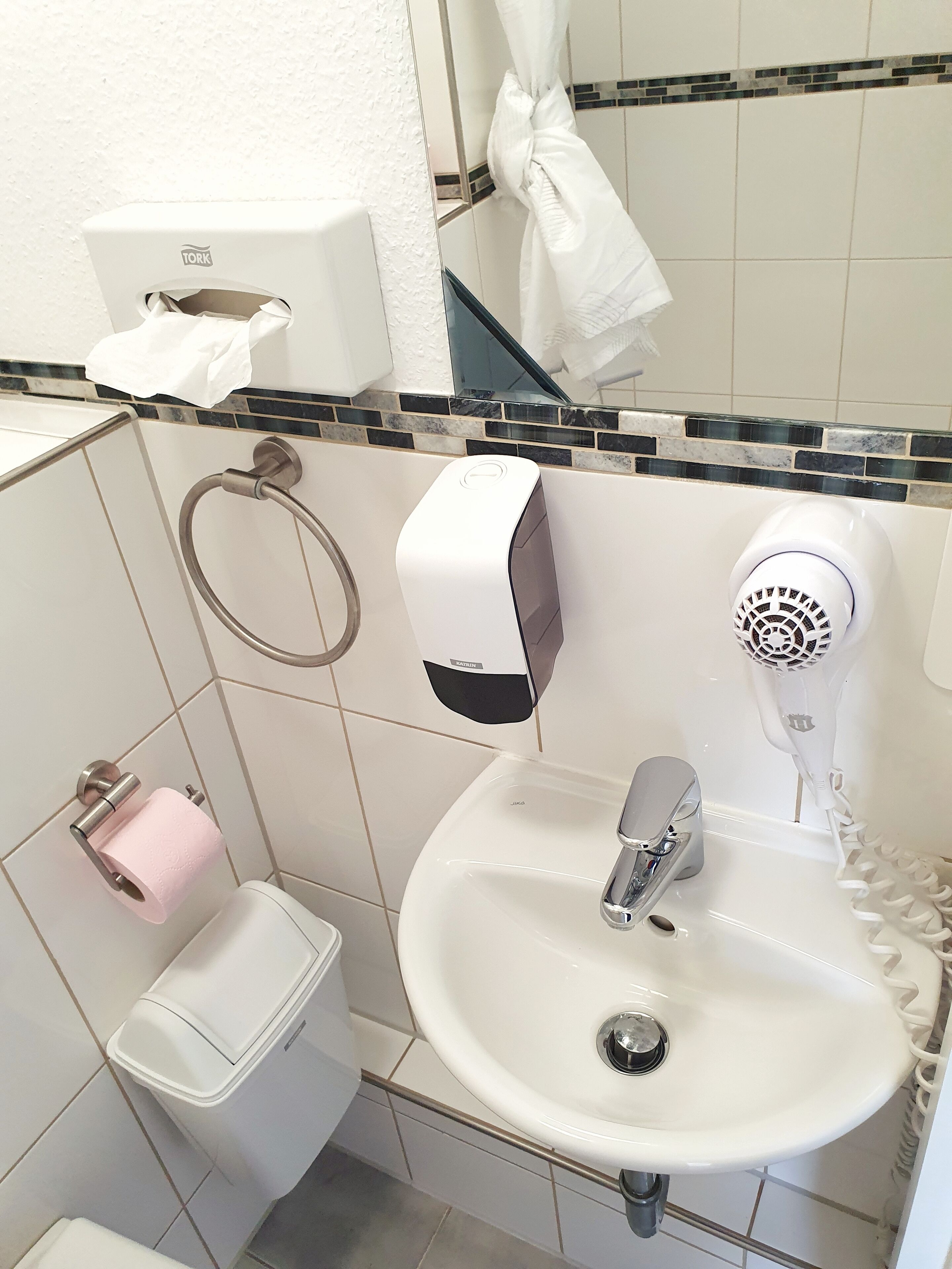 Casa de banho com lavatório branco, dispensador de papel, suporte para papel higiénico e porta-toalhas.