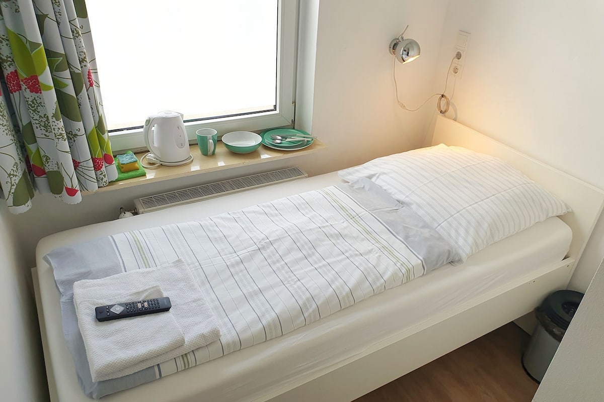 A small, neatly made bed with a remote control on it, a window with curtains, a wall-mounted light, and a small table with a kettle and cups.