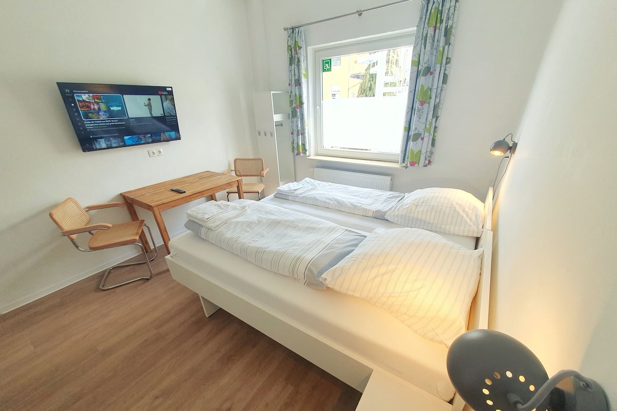 A bedroom with a bed, a desk, a chair, a TV, and a window with curtains.