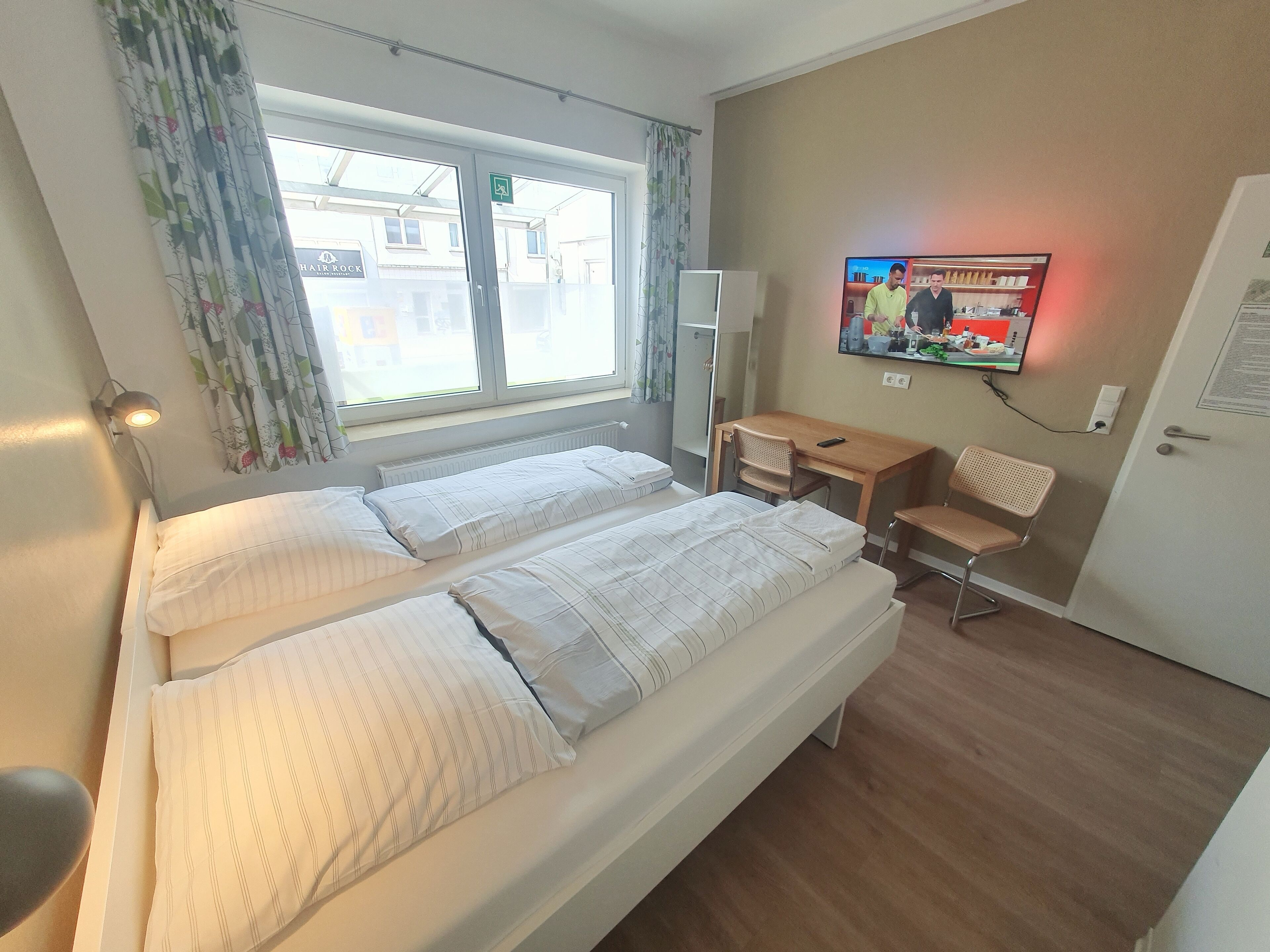 Quarto de hotel com duas camas, uma escrivaninha, uma cadeira, uma televisão e uma janela com cortinas.