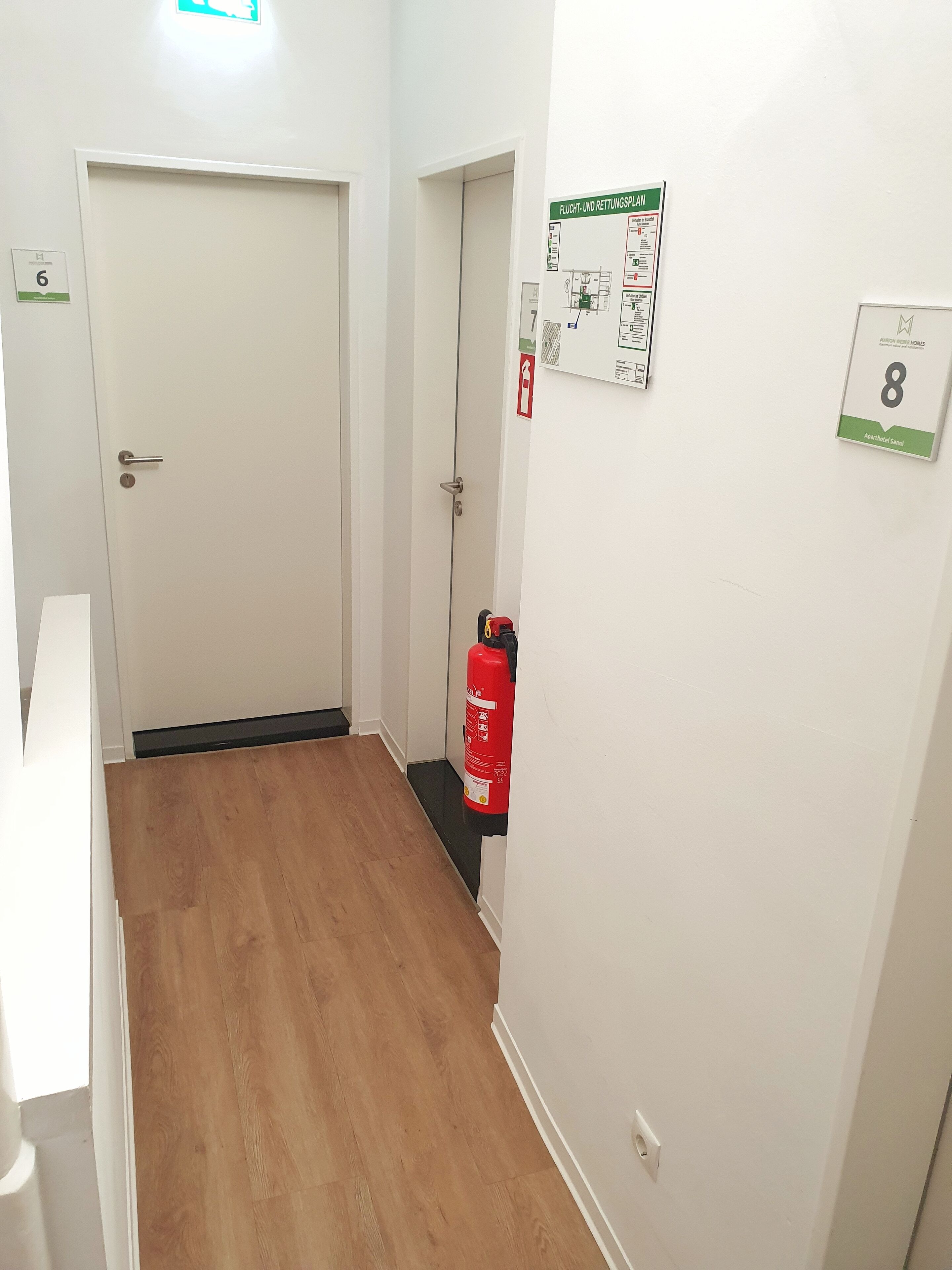 A hallway with a wooden floor, white walls, and a fire extinguisher mounted on the wall.