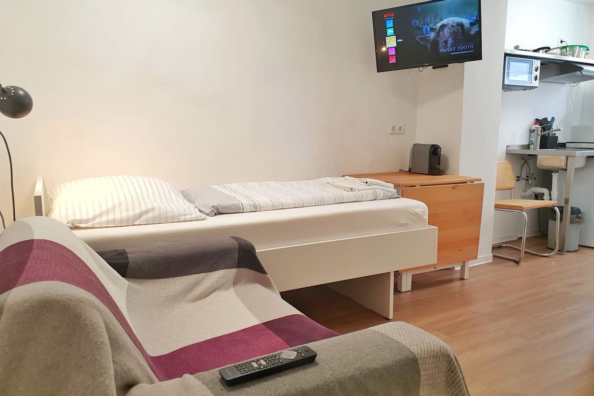 A bedroom with a bed, a TV mounted on the wall, a desk with a chair, and a kitchenette area.
