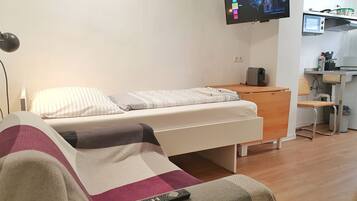 Ein Schlafzimmer mit Bett, Wandfernseher, Schreibtisch mit Stuhl und einer Küchenzeile.