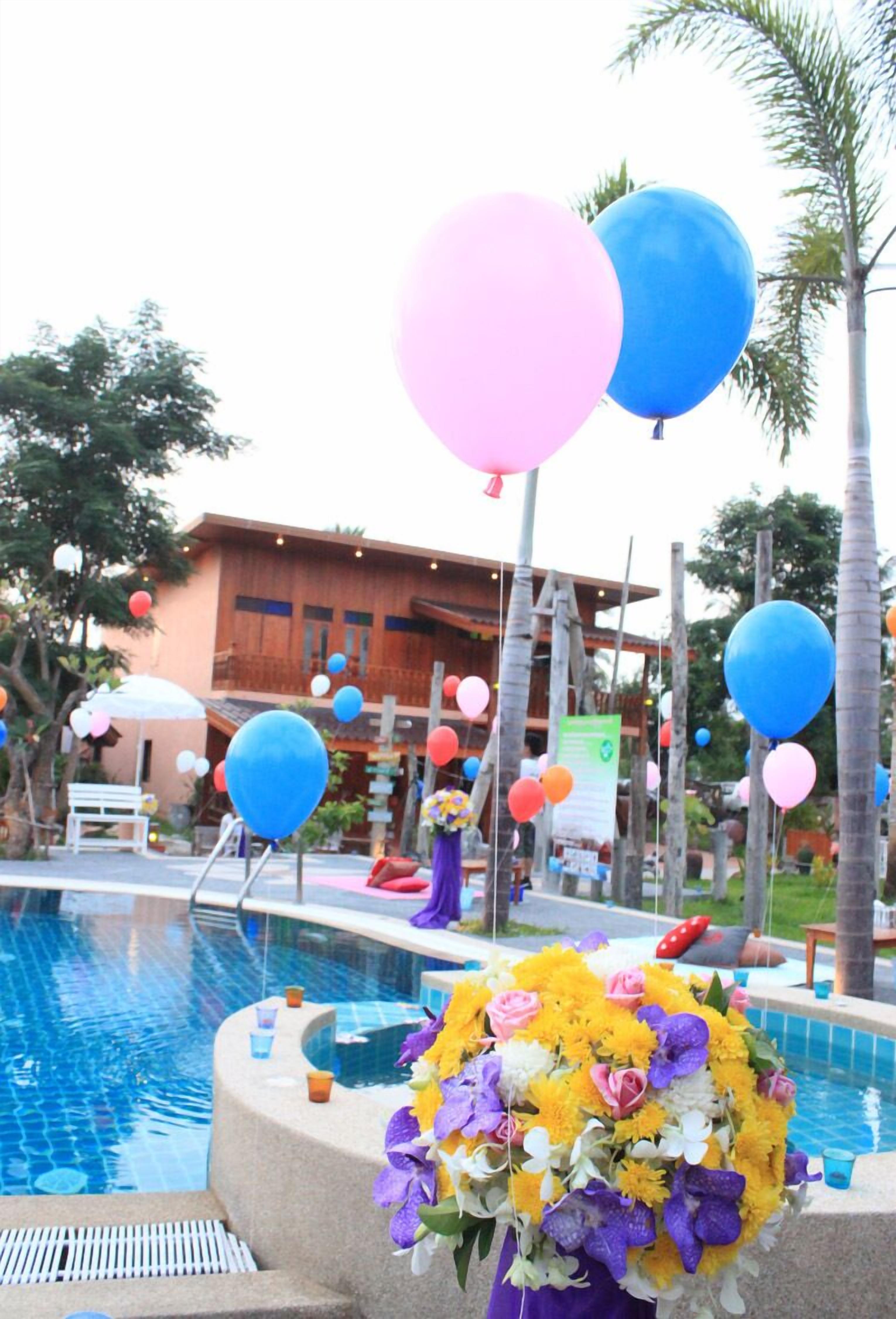 Un área de piscina con un edificio de madera al fondo, rodeada de palmeras y decorada con globos.