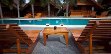 Terraza de madera con sillones de descanso y una mesa, con vista a una piscina.