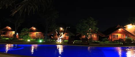 Vista nocturna de un complejo turístico con varias cabañas de madera, una piscina y un jardín bien iluminado.