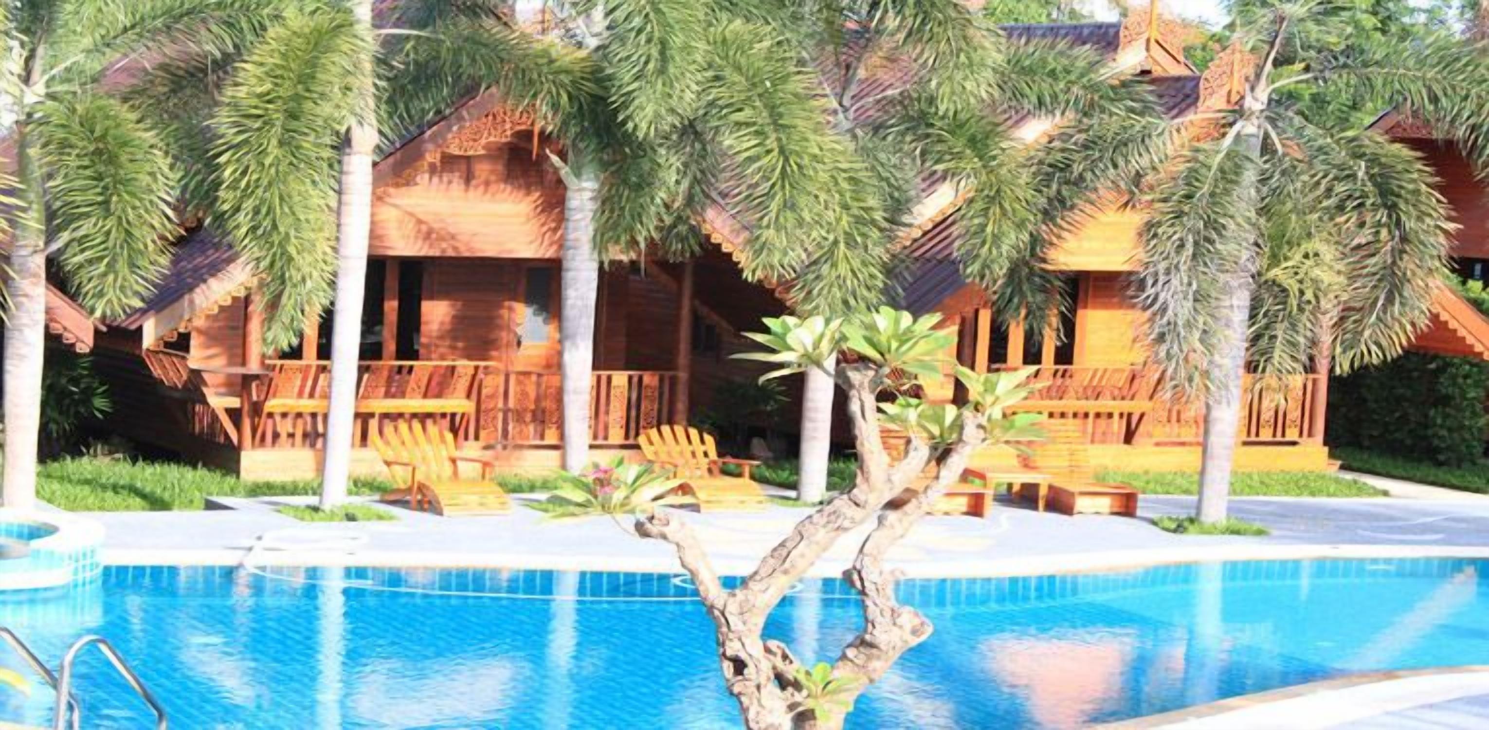 Un área junto a la piscina con una terraza de madera, sillones de descanso y una casa tropical con techo de paja.
