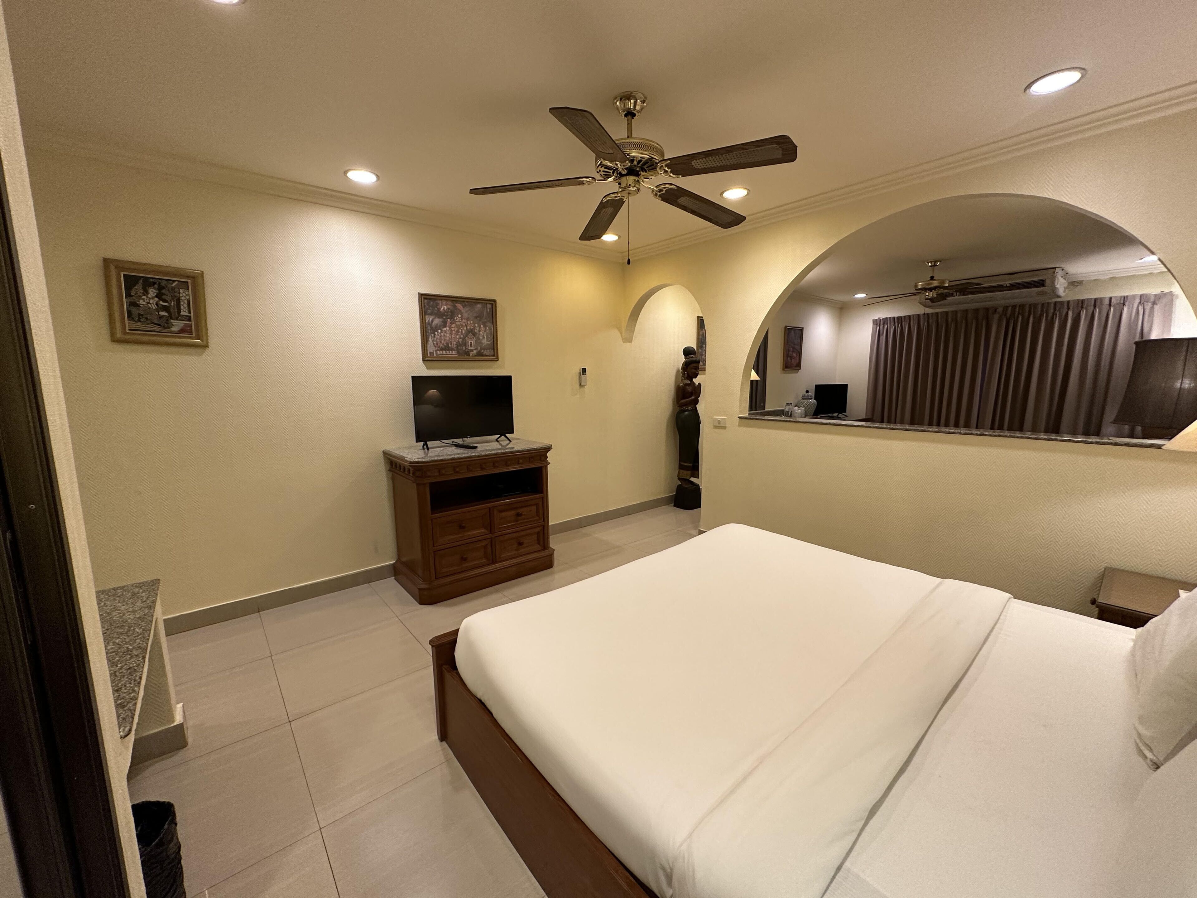 A hotel room with a bed, a TV on a wooden stand, a ceiling fan, and a bathroom with a mirror and curtains.