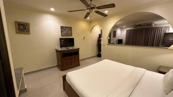 Una habitación de hotel con una cama, un televisor sobre un mueble de madera, un ventilador de techo y un baño con espejo y cortinas.