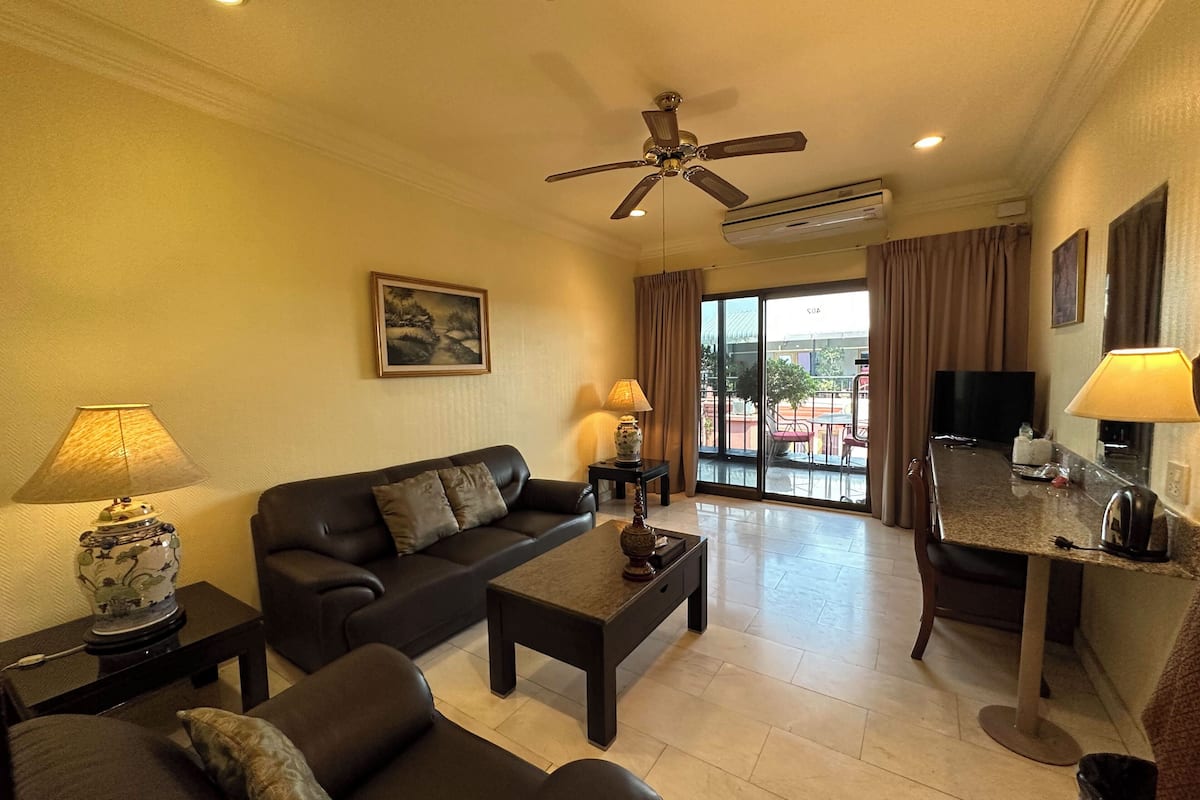 A hotel room with a sofa, coffee table, and a dining area with a table and chairs. There is a ceiling fan and an air conditioning unit.