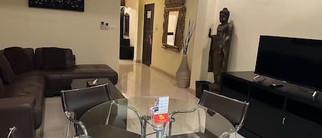 A modern living room with a glass-top dining table, a brown leather sofa, a TV on a black stand, and a statue on the wall.