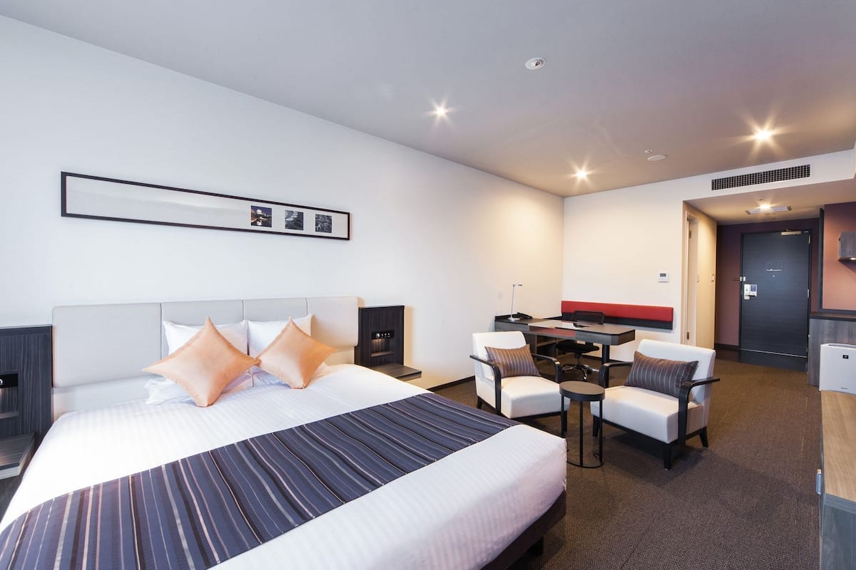 Quarto de hotel com cama, escrivaninha, duas cadeiras, uma mesinha e área de cozinha compacta.