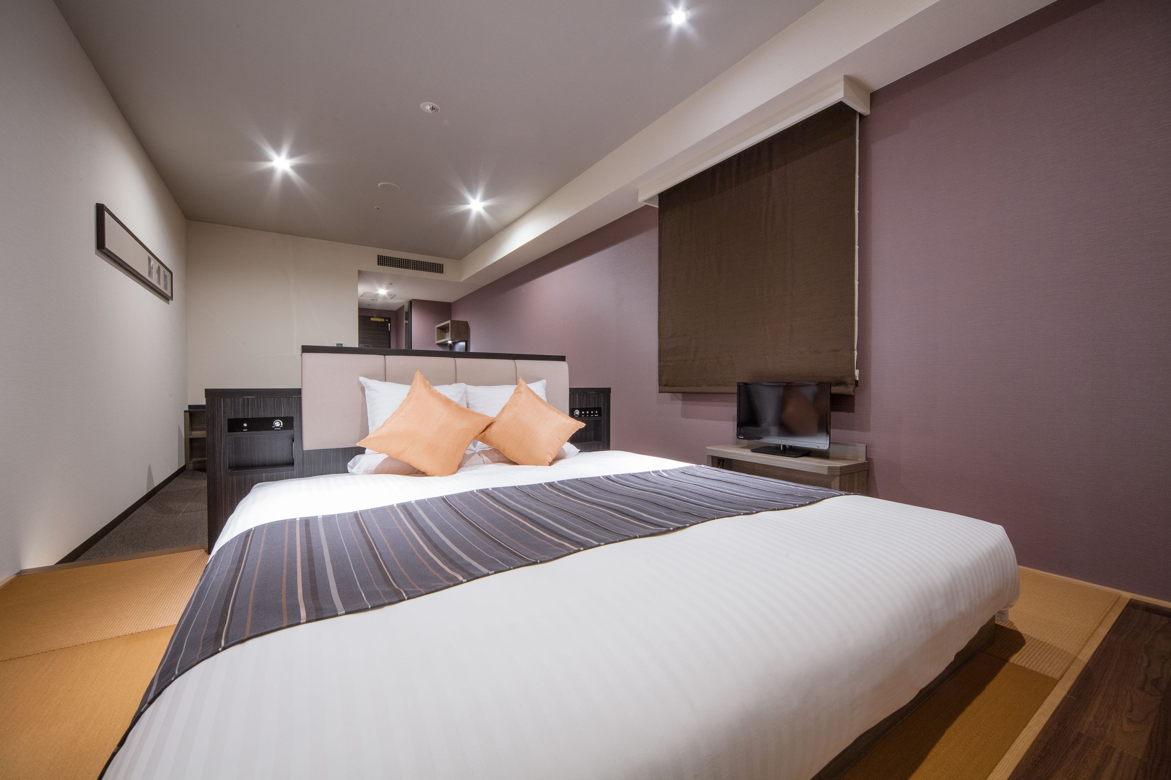 A modern hotel room with a large bed, bedside tables, a flat-screen TV, and a large window with blinds.