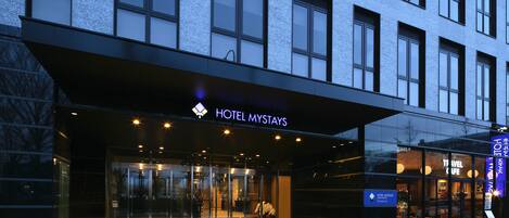 A modern hotel entrance with glass doors, a sign reading "HOTEL MYSTAYS," and a visible reception area.