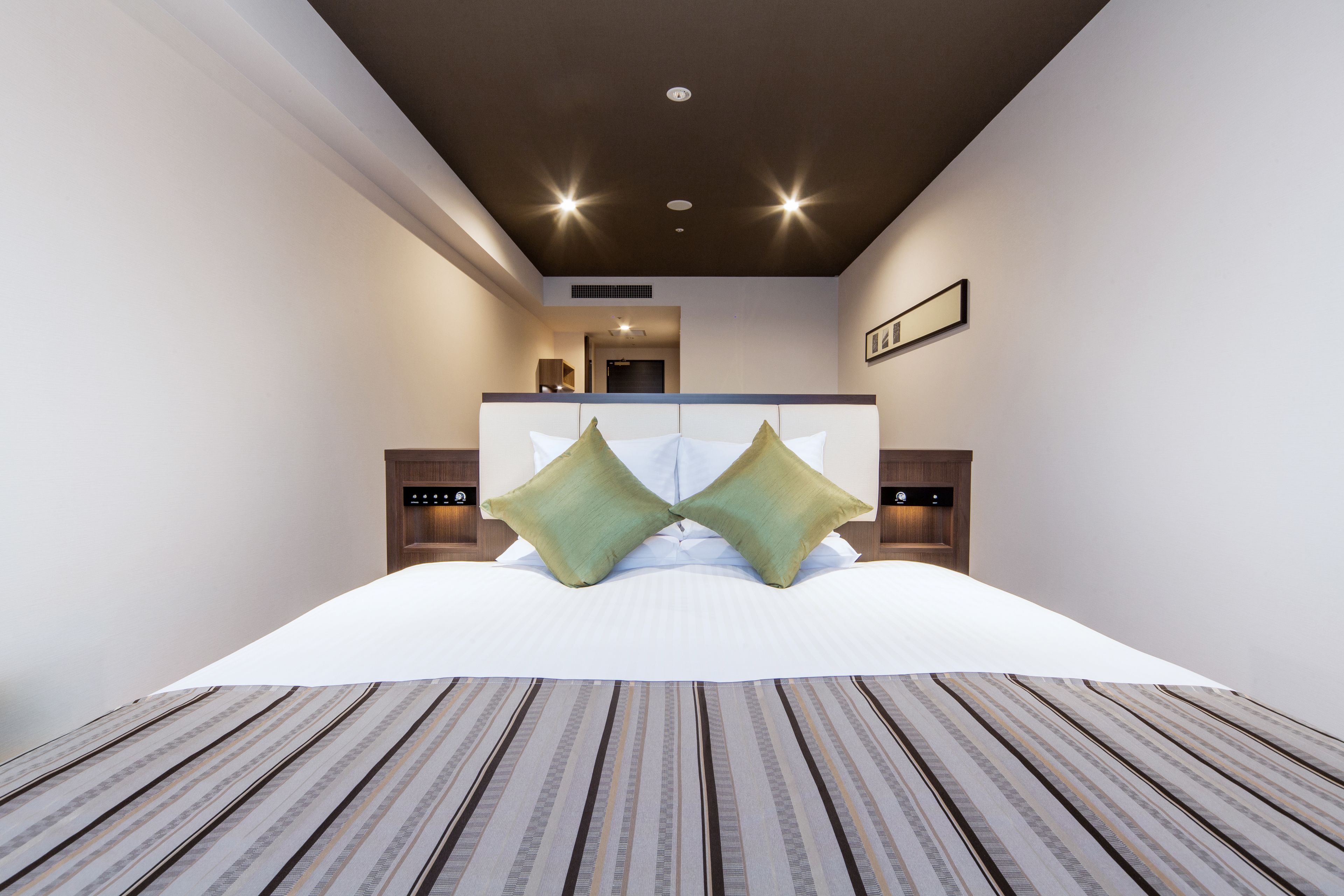 Une chambre d’hôtel moderne avec un grand lit, deux tables de chevet, un luminaire au plafond et un tableau encadré au mur.