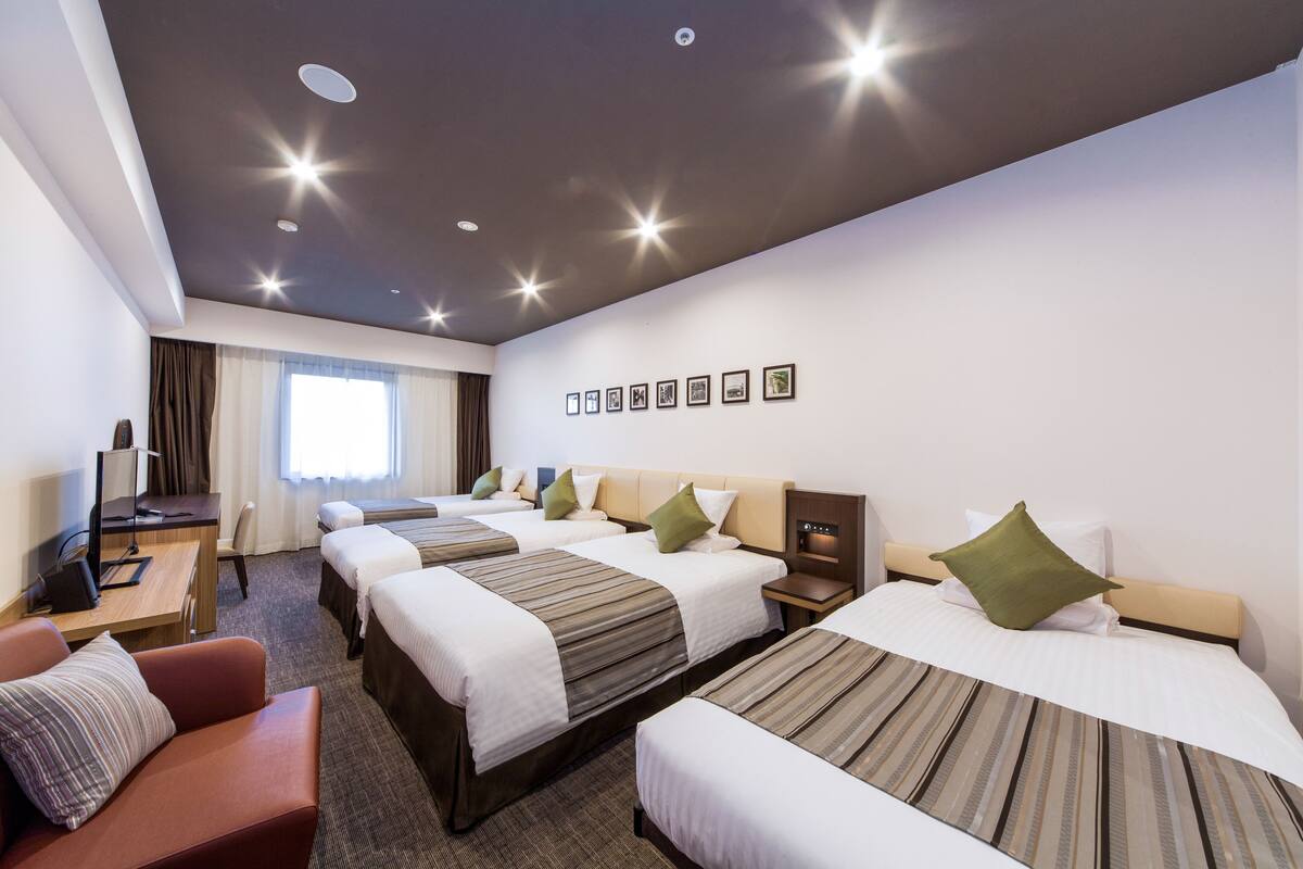 Hotellihuone, jossa on kolme erillistä sänkyä, työpöytä tuolilla, televisio ja ikkuna, jossa on verhot.