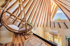 Ein Zimmer mit hohen Decken, einem hängenden Bambusstuhl, einem kleinen Topfpflanzen und Blick ins Freie durch ein Fenster.