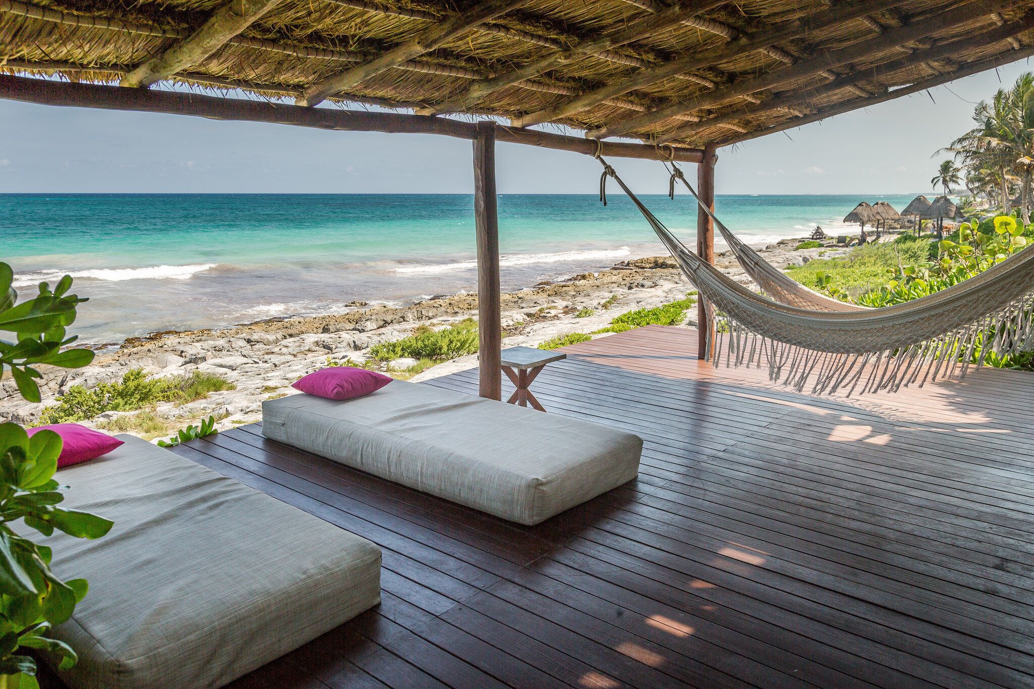 A wooden deck with two cushioned seating areas and a hammock, overlooking a beach with clear turquoise water.