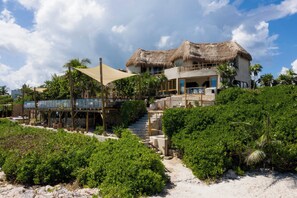Una propiedad frente a la playa con un edificio de techo de paja, un espacio de estar techado al aire libre y abundante vegetación.