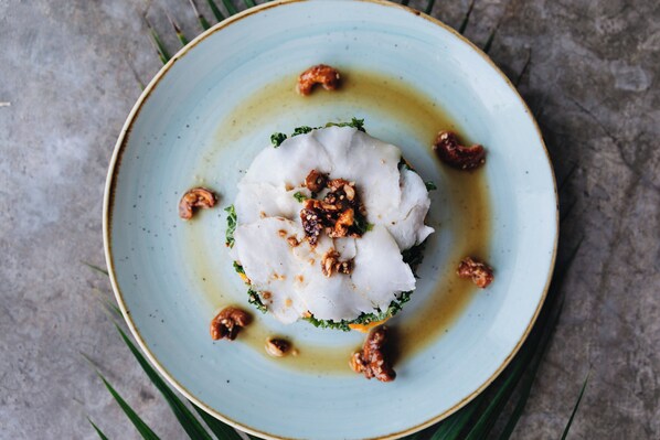 A plate with a white food item, possibly a dumpling, garnished with nuts and herbs, on a blue plate with a light sauce. - Mezzanine by La Zebra, an SLH Hotel (Tulum)