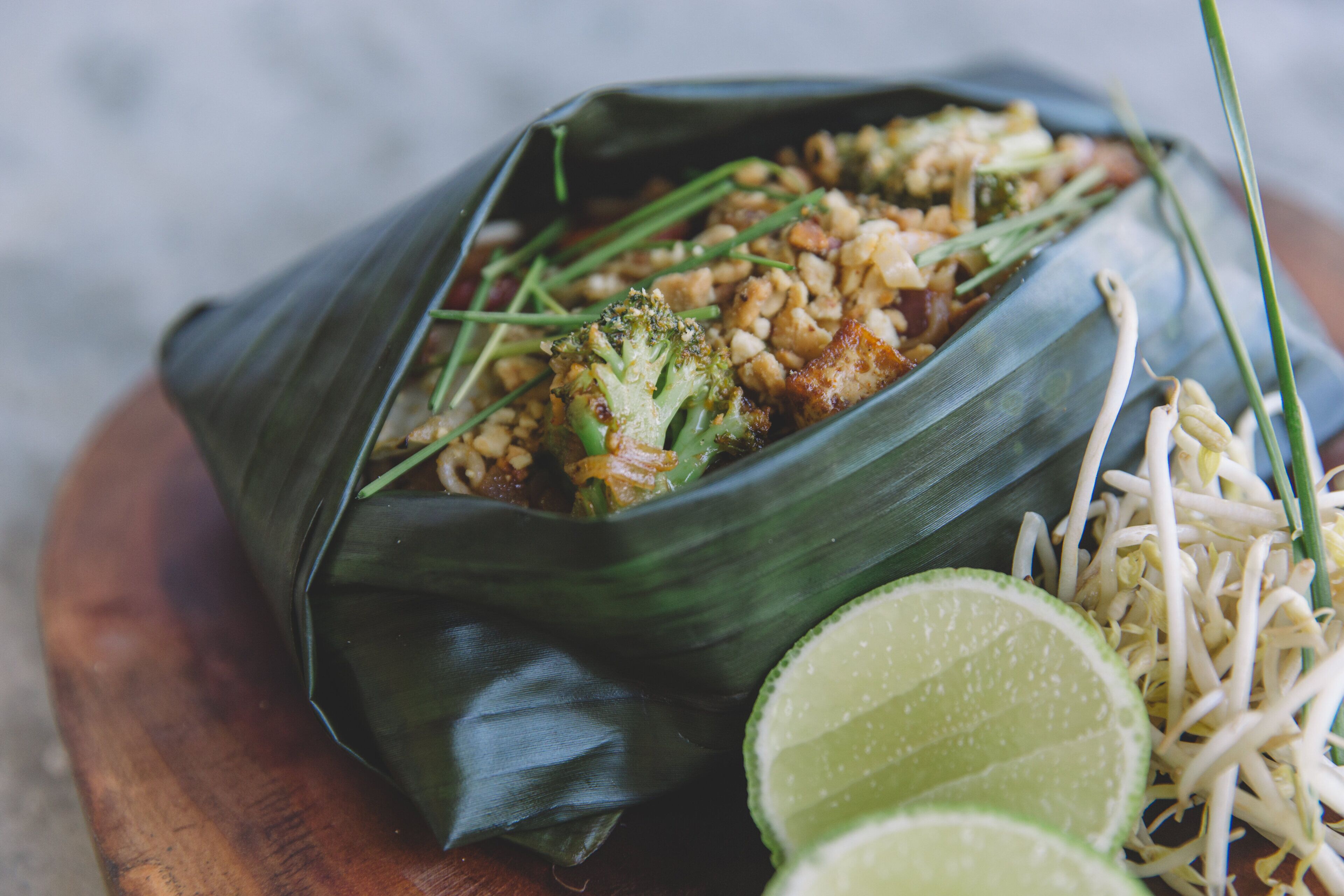 A meal served in a banana leaf bowl with broccoli, rice, and bean sprouts, accompanied by lime wedges.