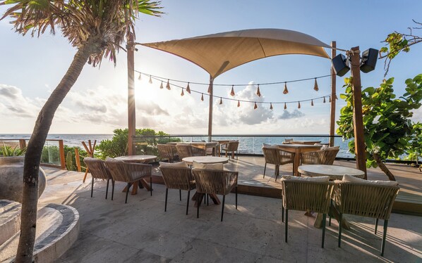 An outdoor seating area with tables and chairs, a large umbrella, and a view of the sea.