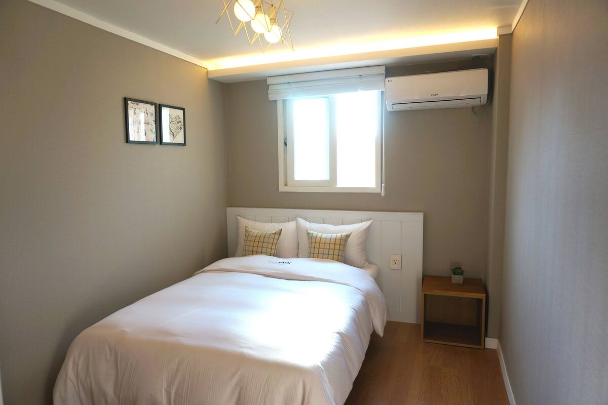 Un dormitorio con cama, ventana, aire acondicionado y una mesita de noche con una planta.