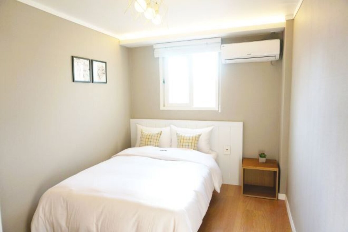 Un dormitorio con cama, ventana, aire acondicionado y una mesita de noche con una planta.