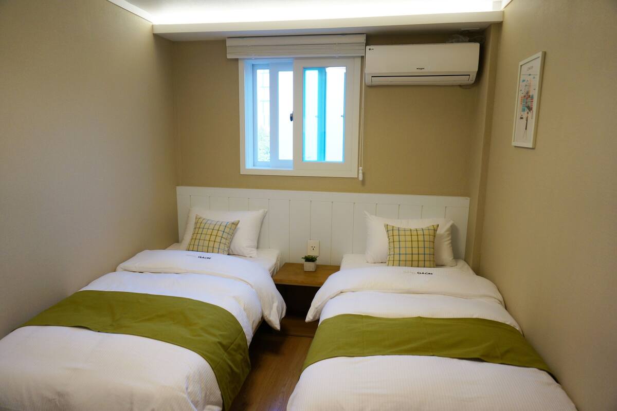Una habitación con dos camas, una ventana con persianas, un aire acondicionado y una pequeña mesa con una planta.