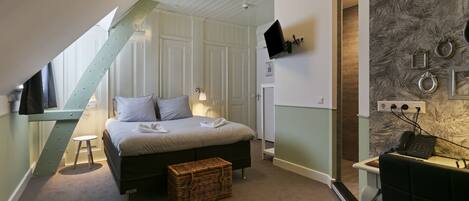 A bedroom with a bed, a small stool, a wicker basket, a desk with a chair, and a mounted TV.