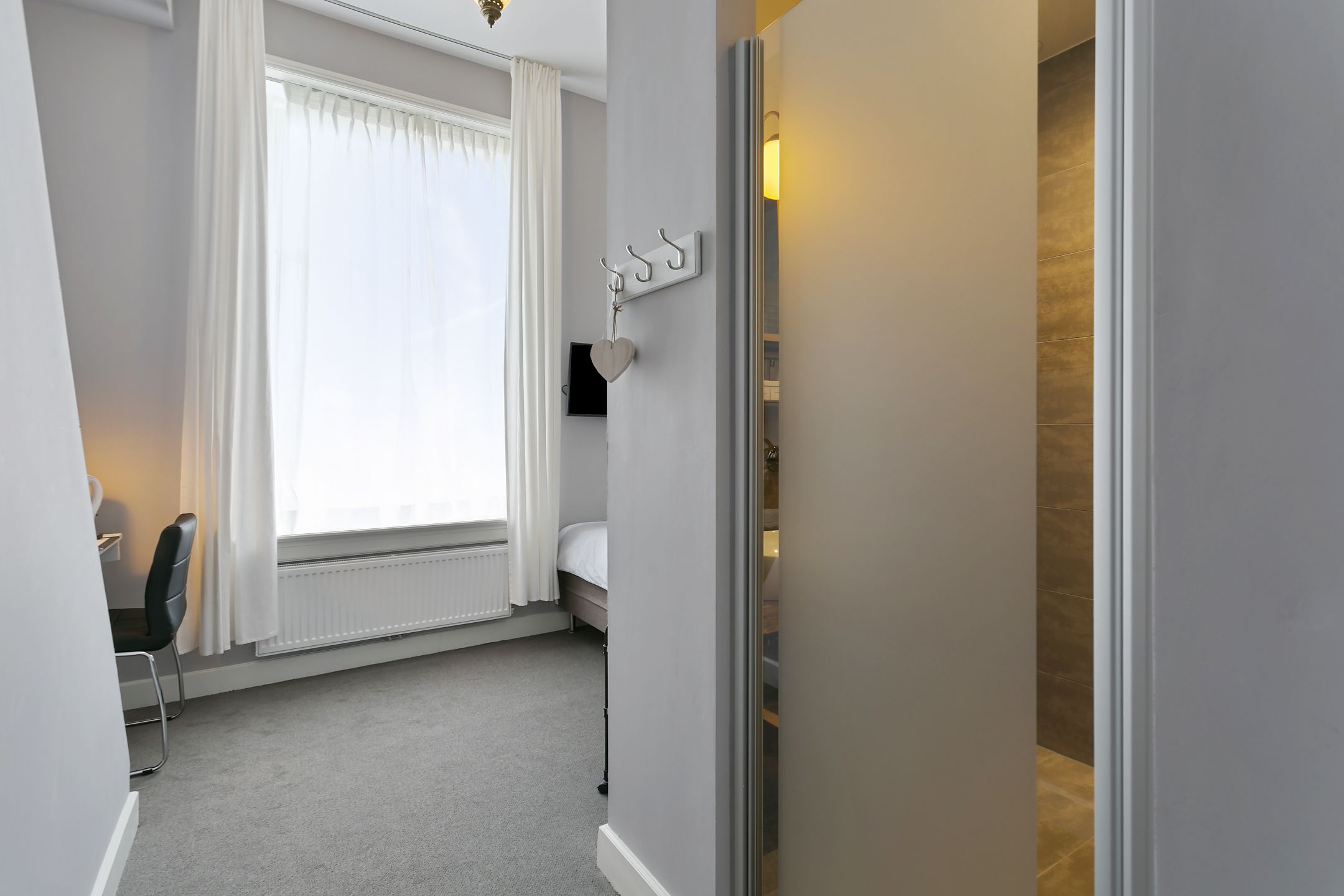 Une chambre d’hôtel avec un lit, une chaise, un radiateur, une fenêtre avec des rideaux et un crochet fixé au mur.