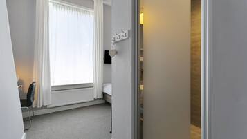 Une chambre d’hôtel avec un lit, une chaise, un radiateur, une fenêtre avec des rideaux et un crochet fixé au mur.